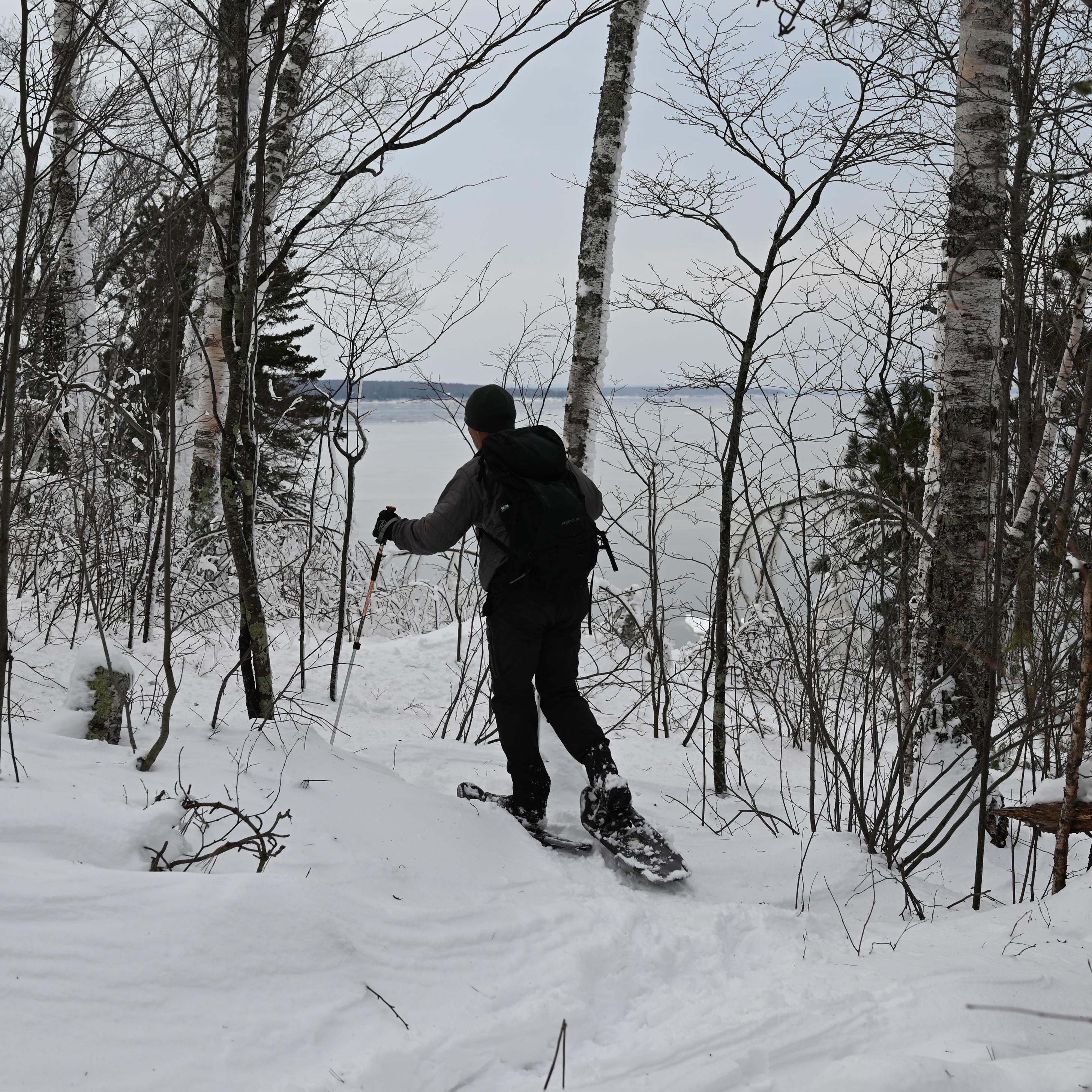 A hiker wearing a grey jacket and snowshoes walks along a snow-covered trail looking over the lake.