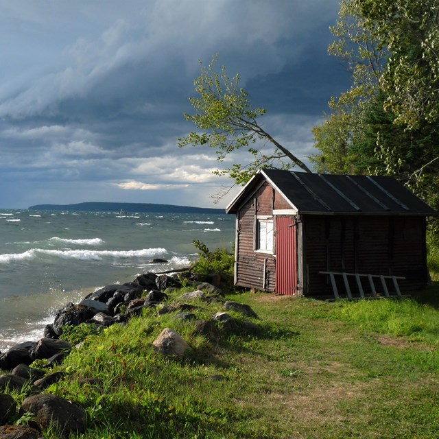 Storm clouds are visible in the distance as waves crash against a rocky shoreline near a small shed.