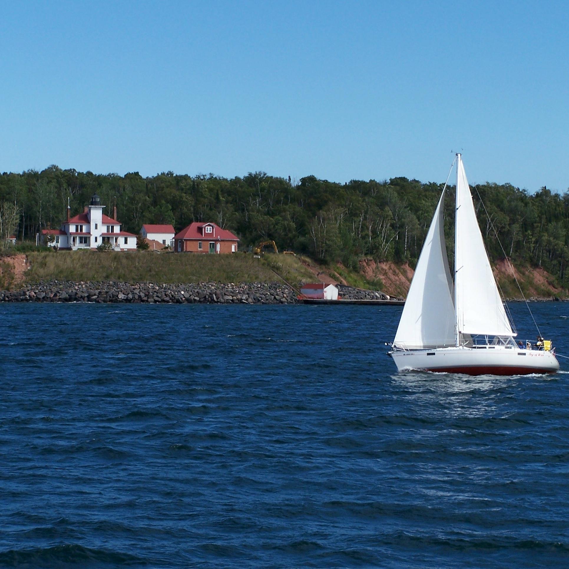 A sailboat cruises past an island with a white lighthouse up on the bluff.