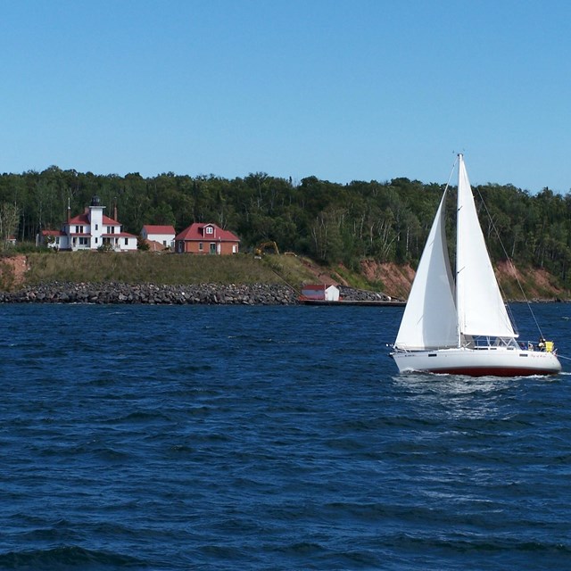 A sailboat cruises past an island with a white lighthouse up on the bluff.