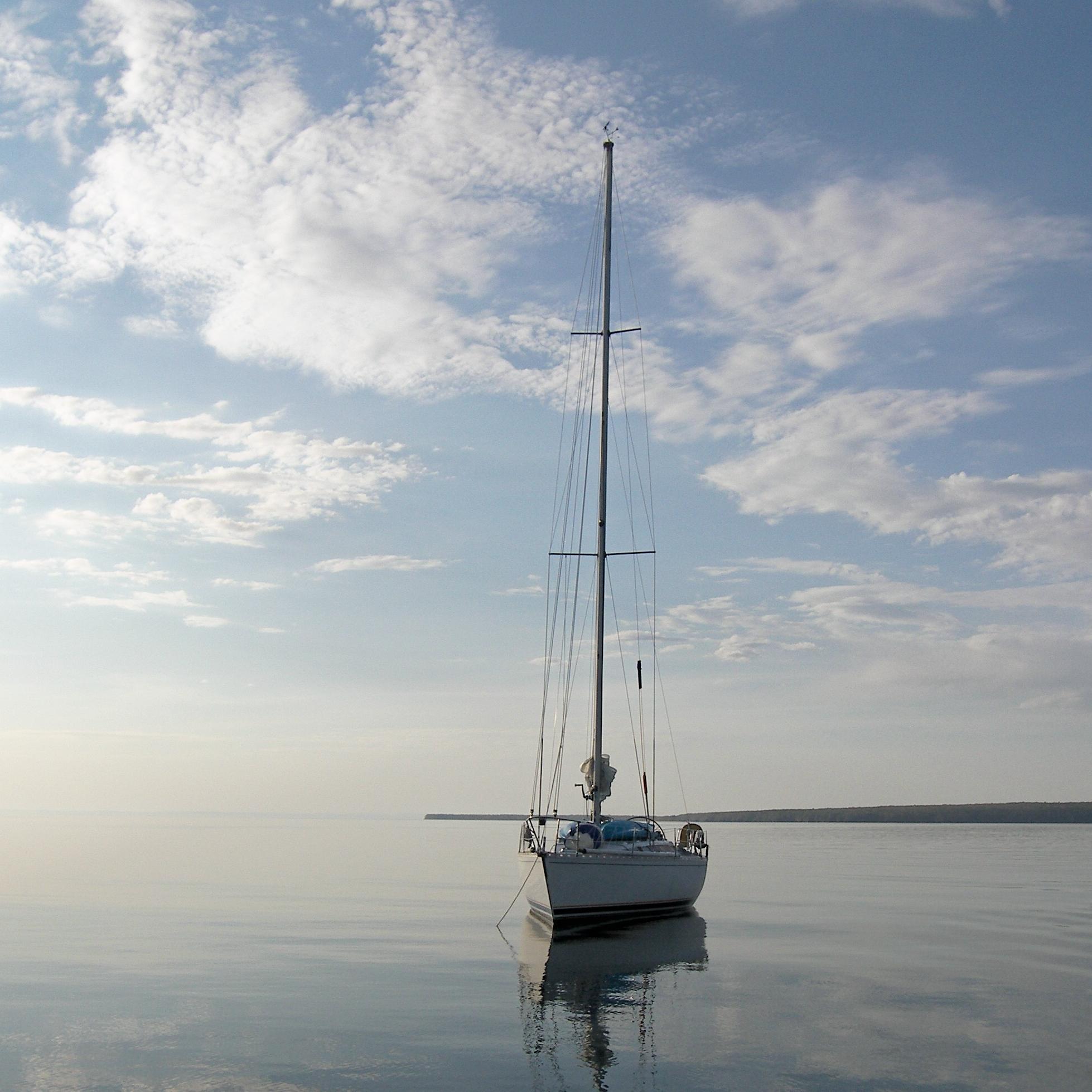 Sailboat with sails down resting on calm water with the sky and clouds reflecting off the surface. 