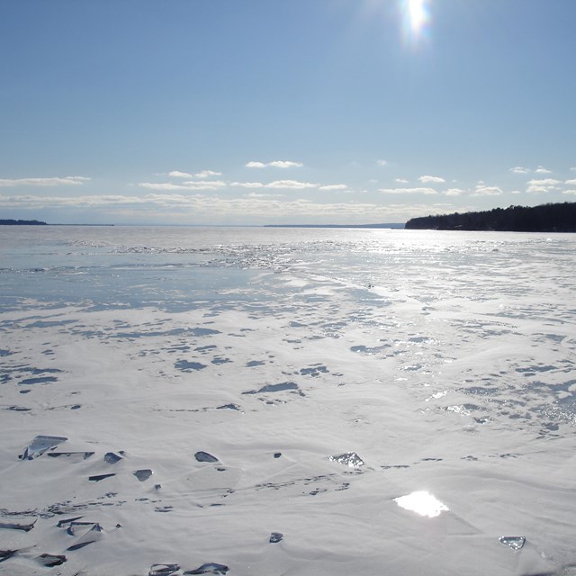 Partially frozen lake with snow drifting across, an island and sun in the background.