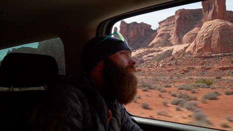 a person with a beard stares out the window at scenery from inside a car. 