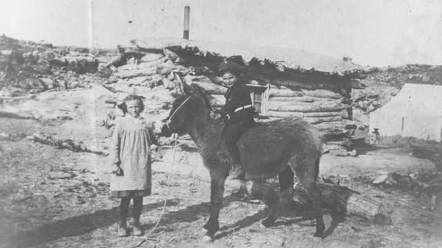 Little girl holding donkey with a boy on its back in front of historic Wolfe Cabin.