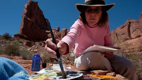 a woman dips a paint brush into a palette with paints