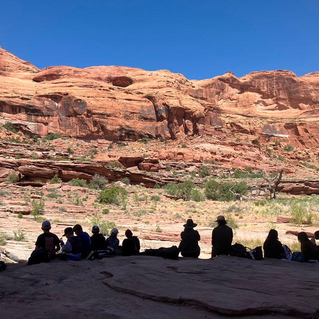 A group of silhouetted children and adults sit on a sandstone bench within a canyon.