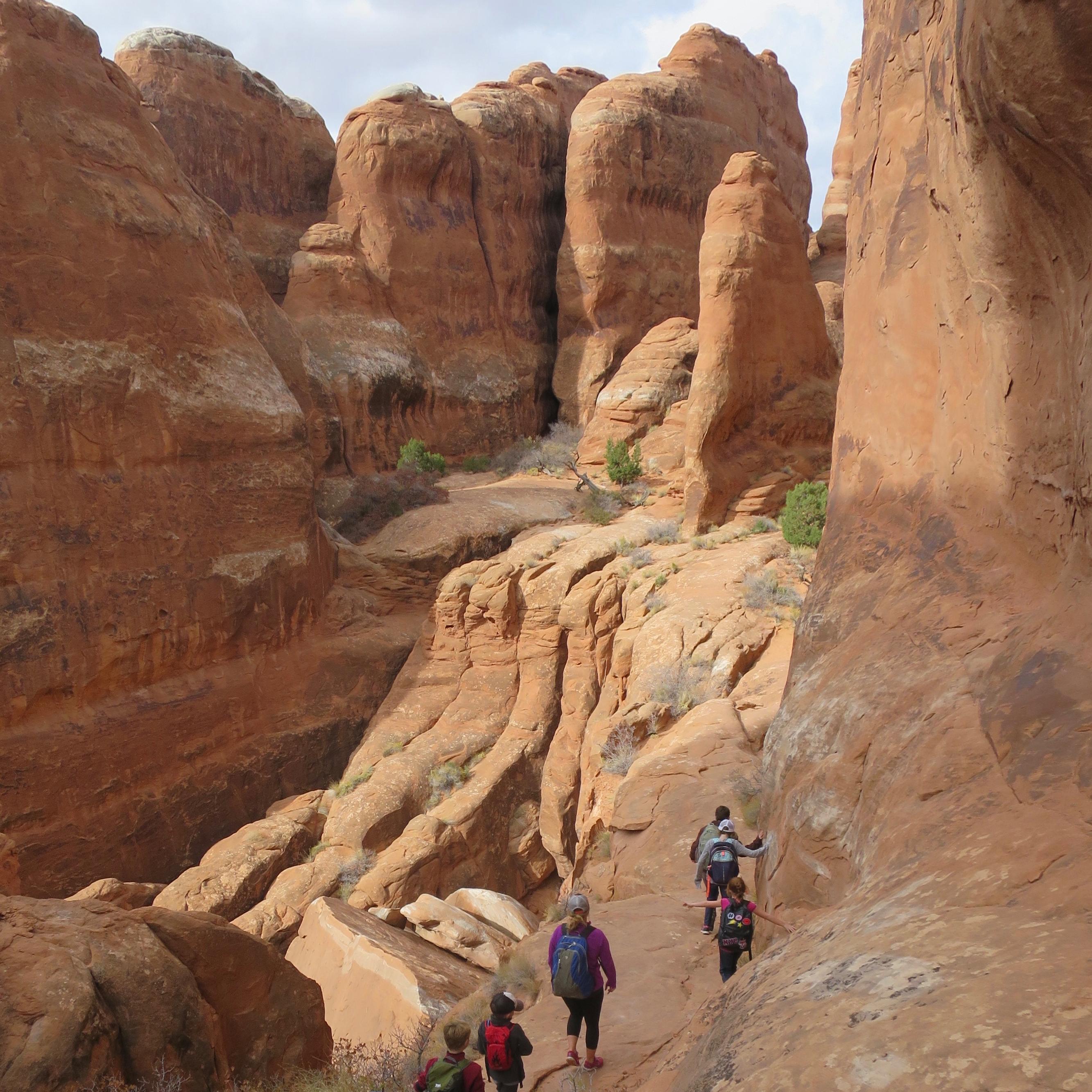 A group of children walk along a narrow section of red sandstone between tall spires and walls.