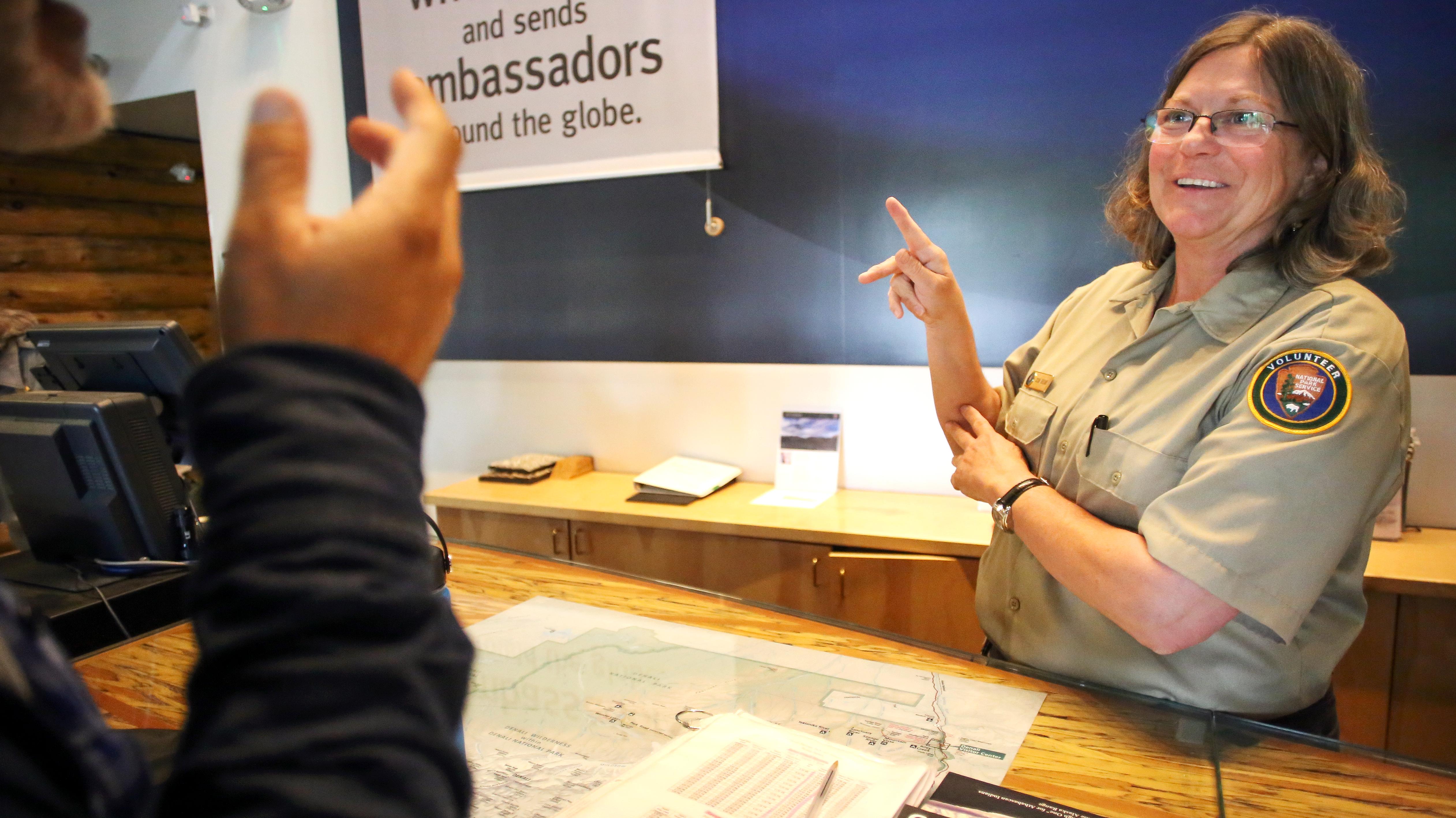 A park volunteer behind a desk communicates with a visitor using sign language.