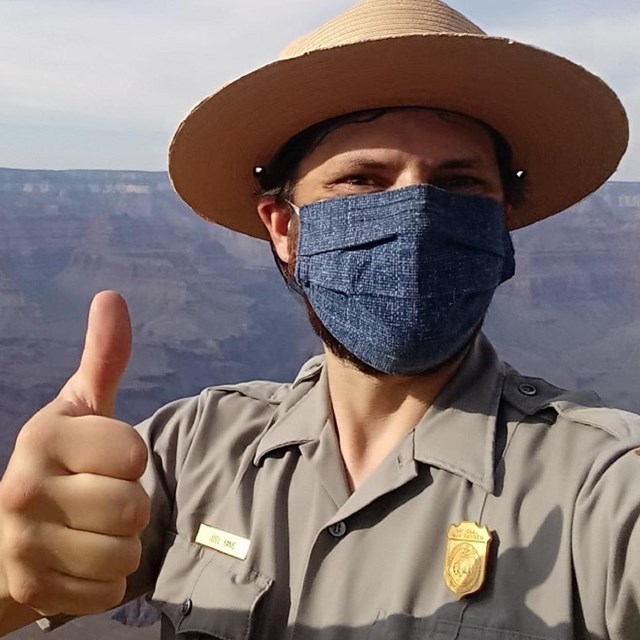 A person giving a thumbs up in ranger uniform and mask, in front of the Grand Canyon