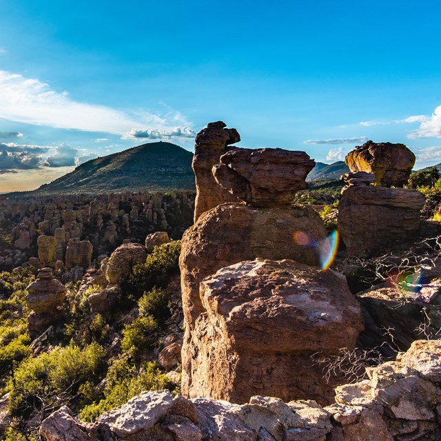 Sunburst, blue sky, and mountain behind standing rock formations and some green trees.