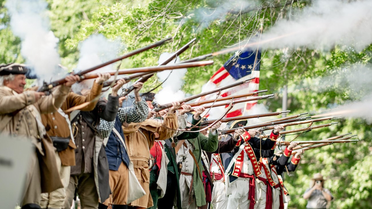 A row of men dressed in colonial militia outfits firings muskets.