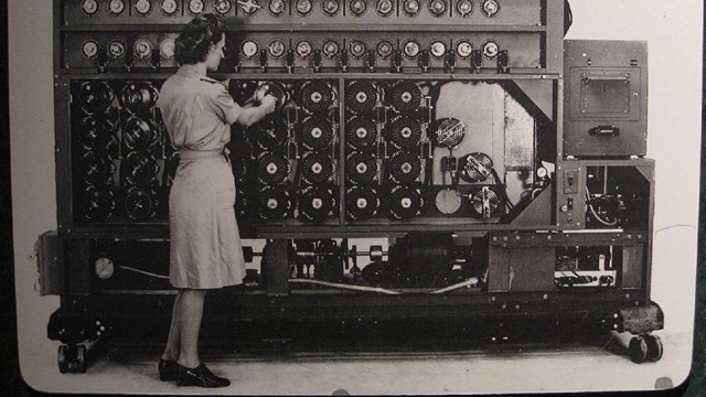 A woman stands in front of a wall of gears and machinery