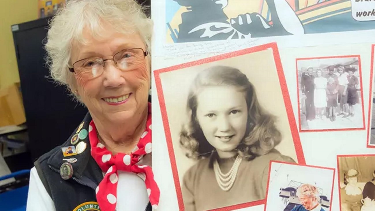 An elderly woman with light skin and white hair holds a poster with historical photos