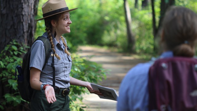 a smiling park ranger talking to an audience on a wooded trail.