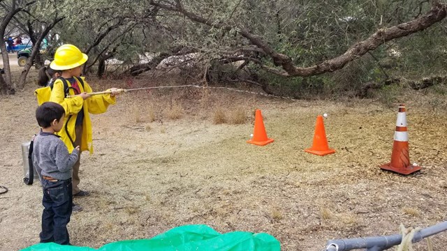 Two children standing near orange cones, one child is spraying water from a fire hose at the cones.