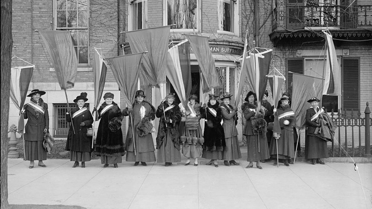 A group of suffragists standing with banner and flags in front of a building. Library of Congress. 