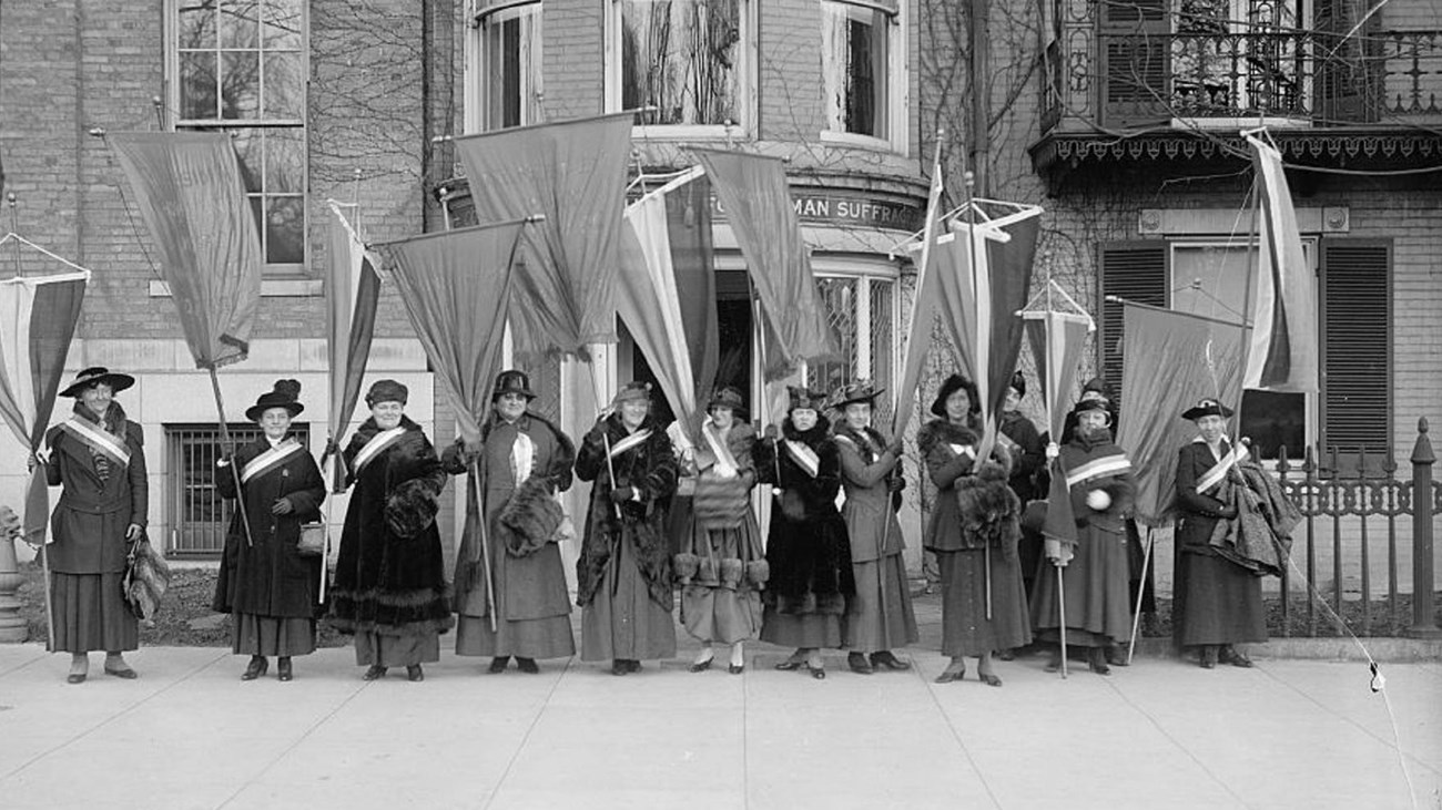 A group of suffragists standing with banner and flags in front of a building. Library of Congress. 