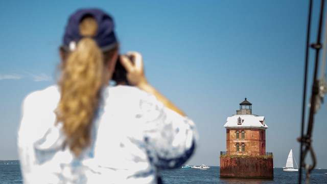 A woman captures a lighthouse with her camera from a boat, surrounded by sailboats.