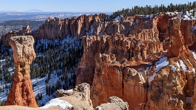 Hoodoos (tall, red stone masses) rise up from the ground and are partially covered with snow.