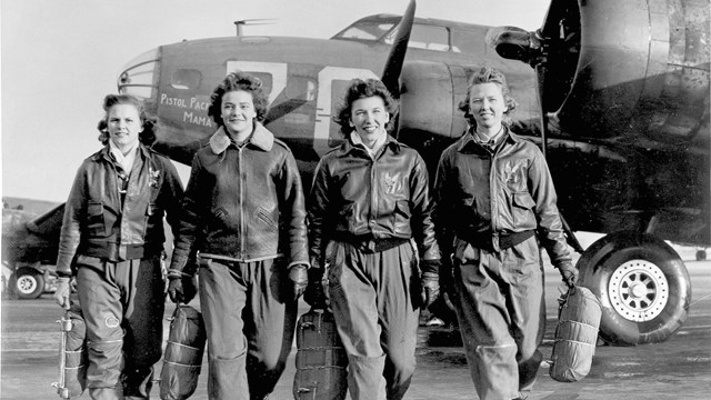 Four women in flight suits walk toward the camera with a plane in the background