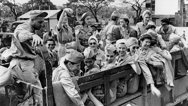 A group of women in military uniform smiles and waves while seated in an open wagon