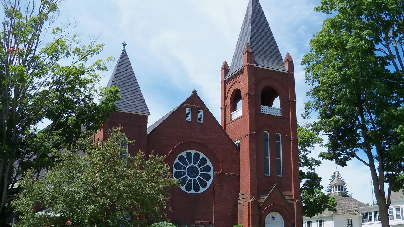 Exterior of red-brick church with steeple & stained-glass window.Photo-by:AlexiusHoratius, CCBYSA3.0