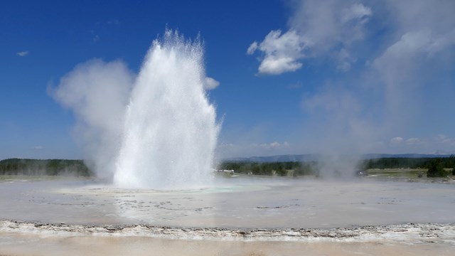 A geyser erupts steam and water out of the middle of a large pool.