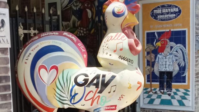 A white rooster statue with colorful decorations and “Gay Ocho” written on its side stands upon a bo