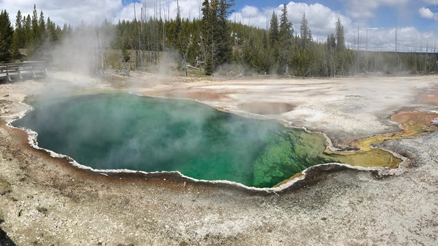 A large, deep blue pool of naturally hot water is surrounded by a rim of white, black, and orange.