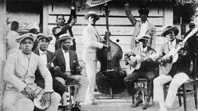 Group of men musicians pose with instruments in front of a house.