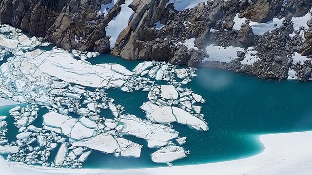 Looking down at a pool of blue water, broken icebergs on the surface, surrounded by a steep cliiff