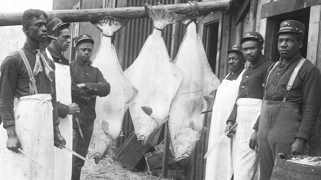 Black and white photo of six troops in Company L standing next to three large halibut fish hanging f