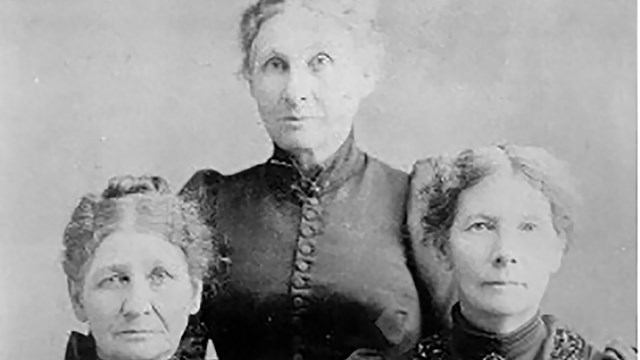 Three women sit for a portrait in 19th century dresses.