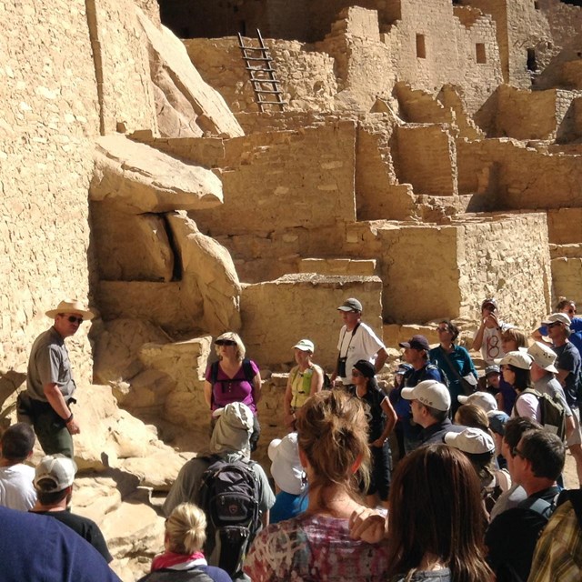 Visitors amass around a ranger with cliff ruins in the background