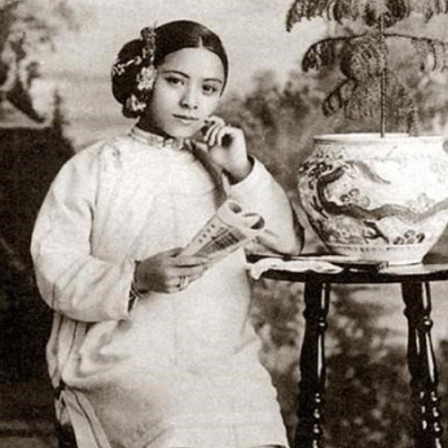 An Asian woman smiling slightly, seated and dressed in white clothes with flowers in her hair