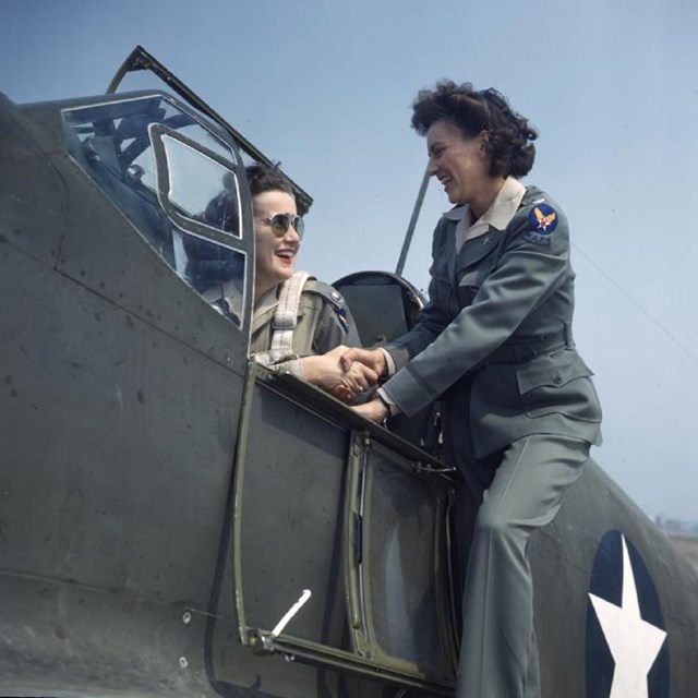 Two white women shake hands and smile; one is sitting in the cockpit of a plane