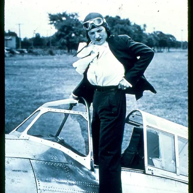 White woman with flying goggles adjusts clothing in cockpit of plane