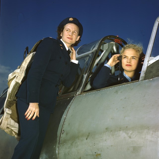 A woman in uniform talks to another woman seated with earmuffs in the cockpit of a plane