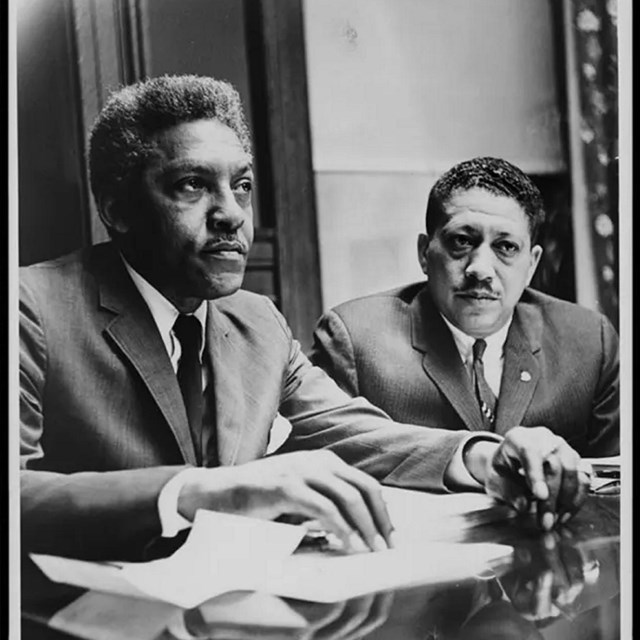 Bayard Rustin, a black man in a suit sitting at a desk next to another man