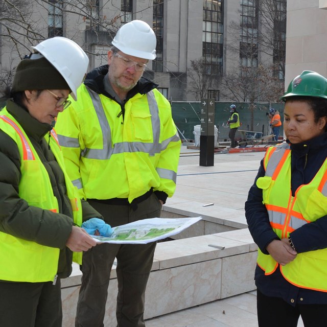 Three staff in hard hats and safety vests looking at plans of a memorial 