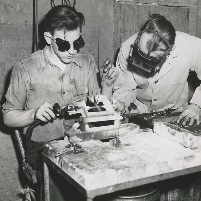 Young man wearing goggles sits at a workbench to weld. Another man inspects his work.