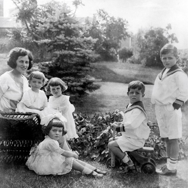 Rose Kennedy and 5 children pose on the lawn. John Kennedy sits on a wooden tricycle.