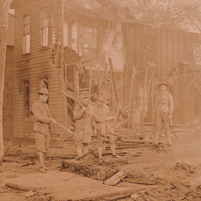Four soldiers stand on the smoking remains of a burned house, with charred framing still standing.