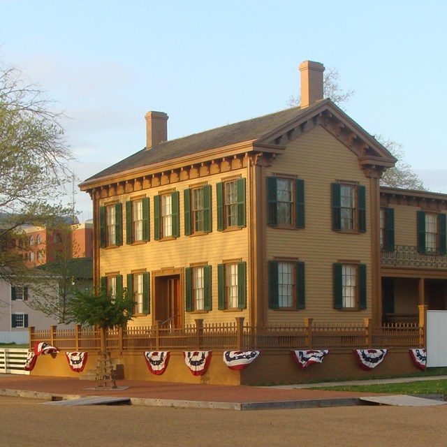 A low brick wall and wooden fence surround a two-story tan house with green window shutters.