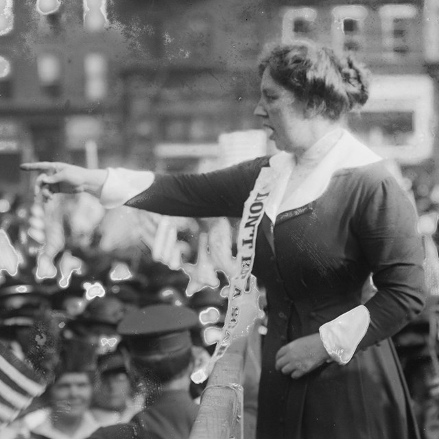 Woman standing in front of a crowd, gesturing with her hand and wearing a white sash