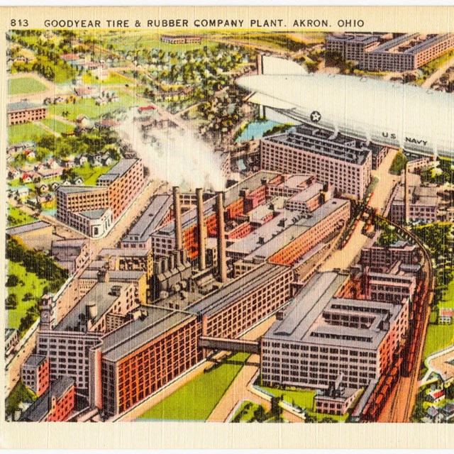 Color postcard of a silver US Navy blimp flying over the Goodyear Tire and Rubber Company plant