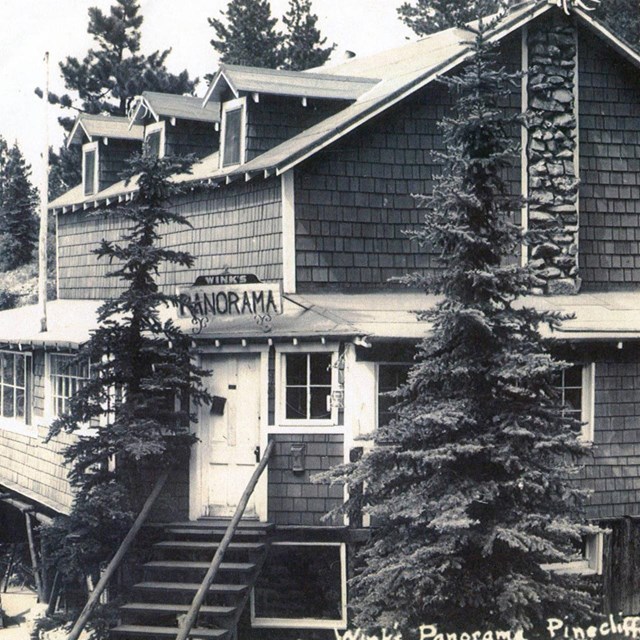 Black and white photo of a two story wooden house in the trees.