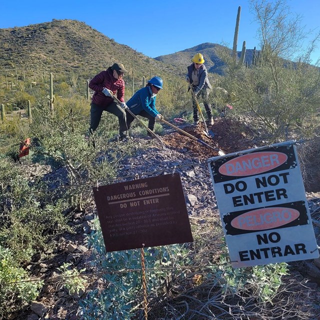 Three young people use shovels behind a fence that says danger do not enter