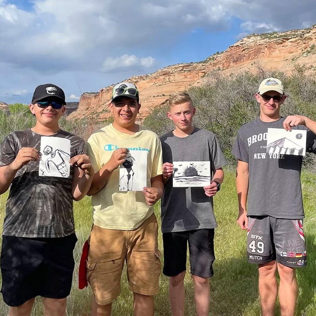 four young men hold up paintings with black ink while standing outside