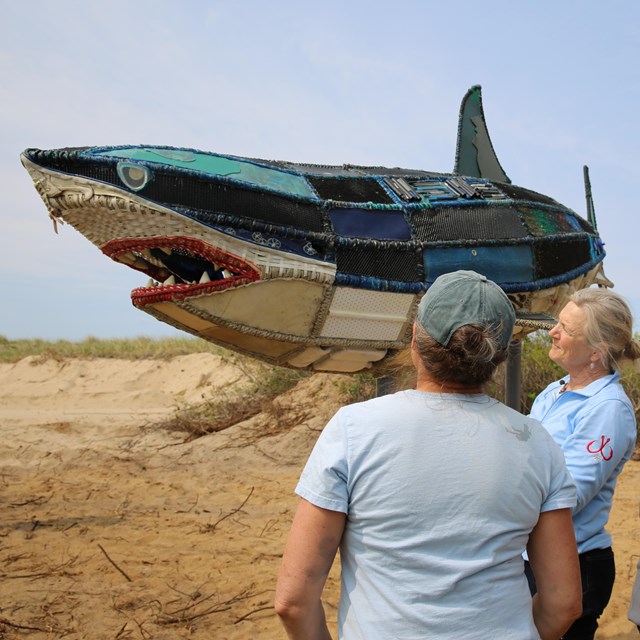 three people next to a 16-foot shark sculpture made of marine debris